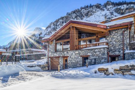 Winter exterior of Chalet Cervinia with stone and timber architecture, large windows, snow-covered surroundings and mountain scenery in bright sunshine.
