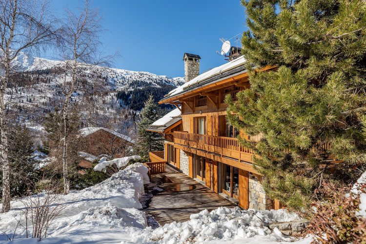 Chalet Foinsbois exterior with a timber balcony, wooden terrace and stone walls, surrounded by snow and trees with mountains in the background.