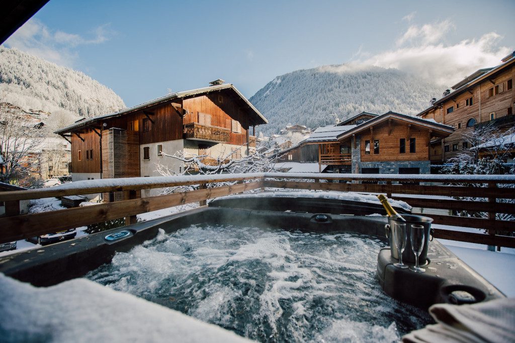 Main outdoor hot tub at Chalet Harmonie with bubbling water, champagne and views across a snow-covered alpine village and forested mountains.