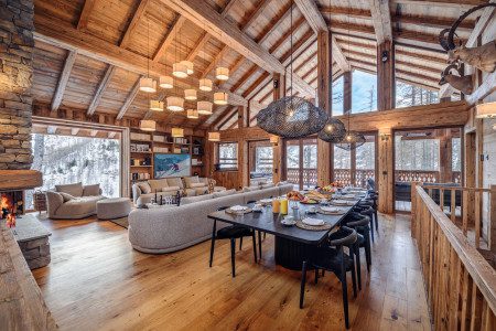 Open-plan living and dining room at Chalet Inoko with a long dining table, soft lounge seating, stone fireplace, exposed timber beams and snowy mountain views.