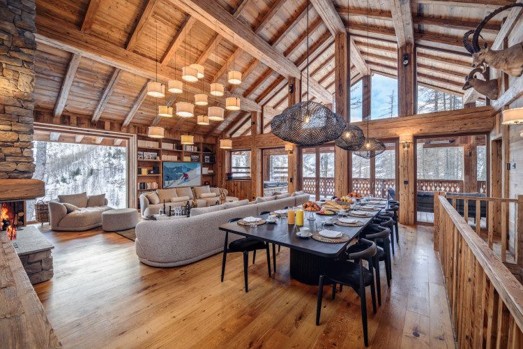 Open-plan living and dining room at Chalet Inoko with a long dining table, soft lounge seating, stone fireplace, exposed timber beams and snowy mountain views.
