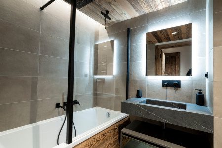 Modern bathroom at Chalet Little Bear with a bath and overhead shower, grey stone tiles, illuminated mirrors and a dark stone sink.