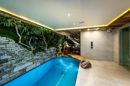 Indoor swimming pool at Chalet Machapuchare with illuminated blue water, natural stone walls, greenery, lounge chairs and modern spa lighting.