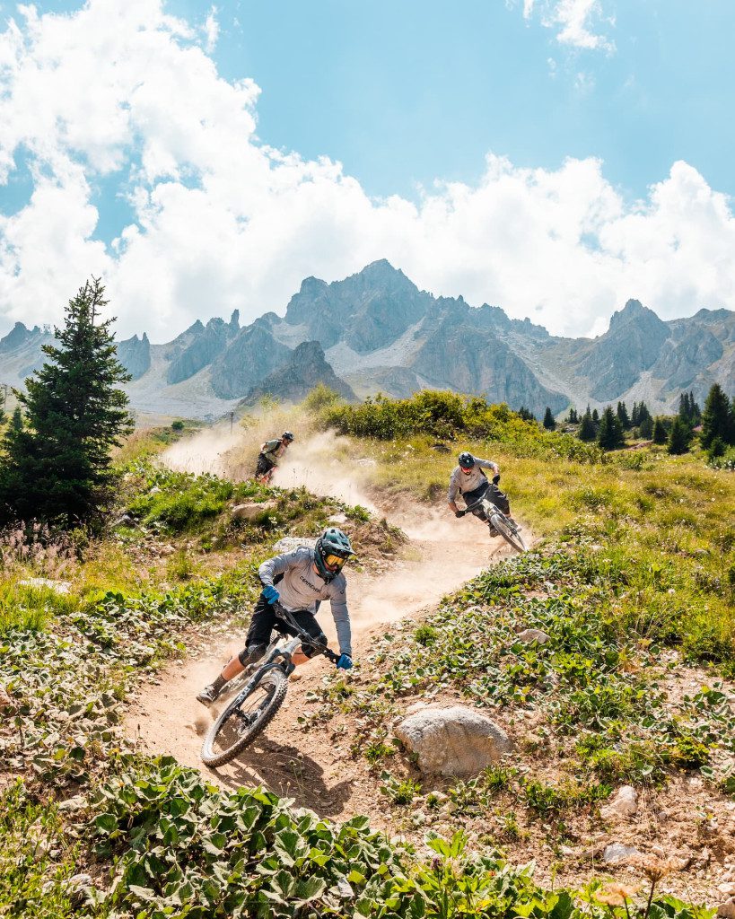 Mountain bikers riding a dusty alpine trail in Courchevel during summer with dramatic rocky peaks in the background.