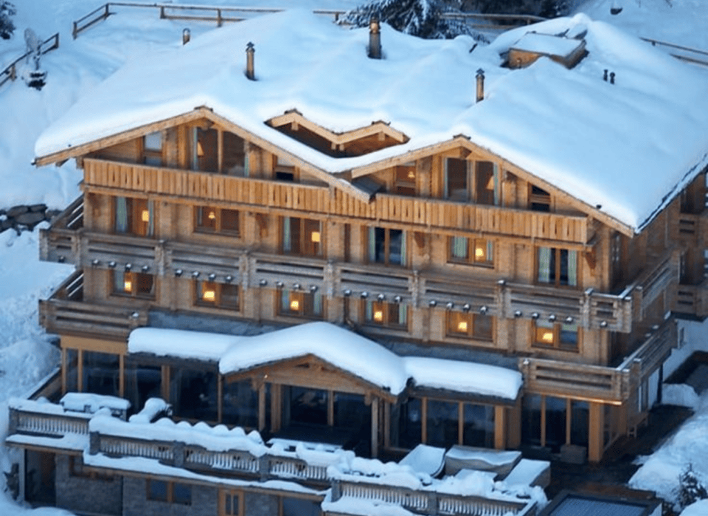 Large luxury ski chalet covered in fresh snow with timber balconies, warm interior lighting and a classic alpine exterior.