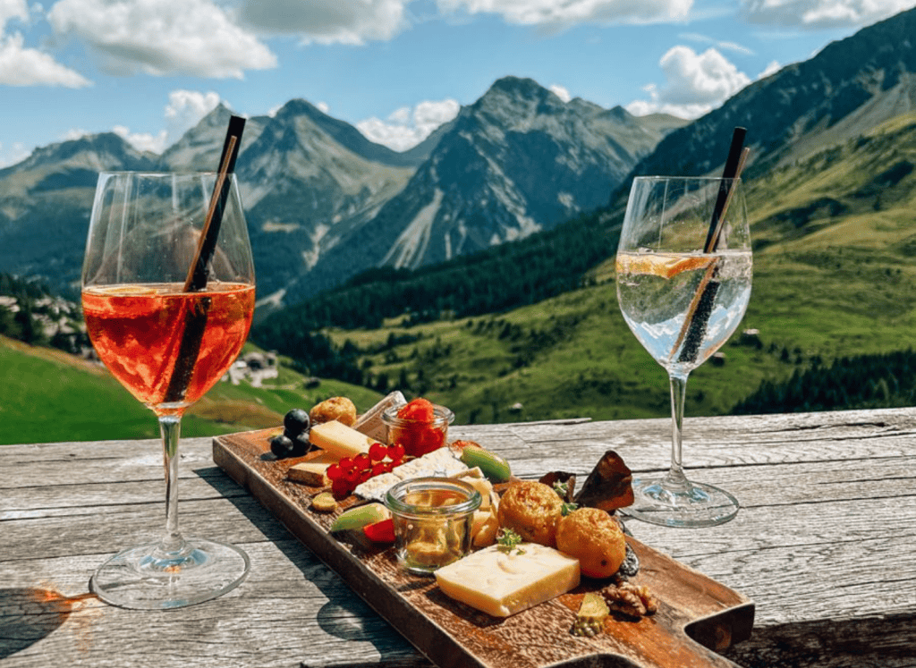 Aperitif drinks and a cheese board on a mountain terrace with panoramic alpine views across green valleys and rugged peaks.