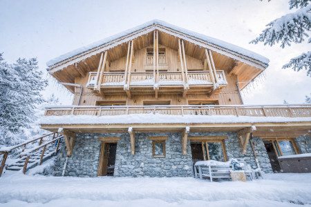 Rear exterior of Chalet La Folie covered in snow with timber balconies, stone walls and snow-covered trees surrounding the chalet.