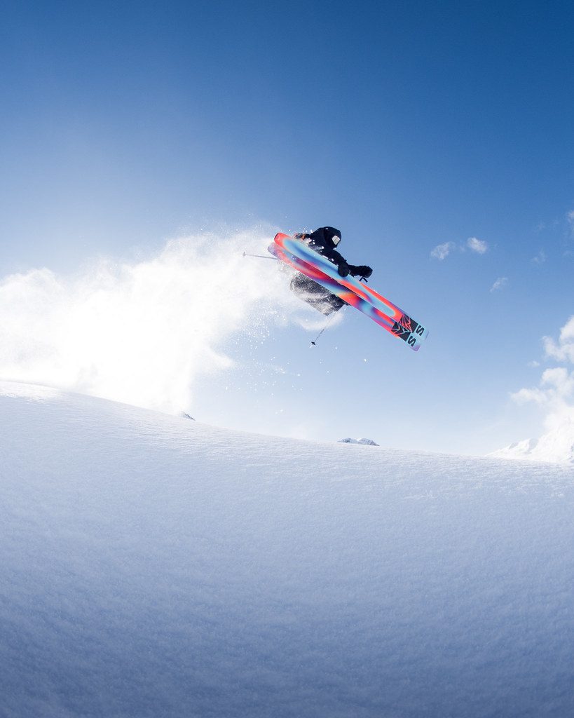 Skier airborne above fresh snow in Sainte Foy Tarentaise, kicking up powder beneath a clear blue sky.
