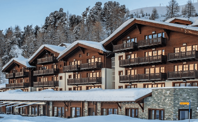 Snow-covered ski accommodation in Switzerland with traditional timber balconies, alpine architecture and forested mountain surroundings.