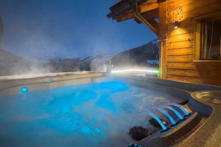 Steaming outdoor hot tub at Apartment La Cascade with illuminated blue water, wooden chalet exterior and snowy mountain views at dusk.