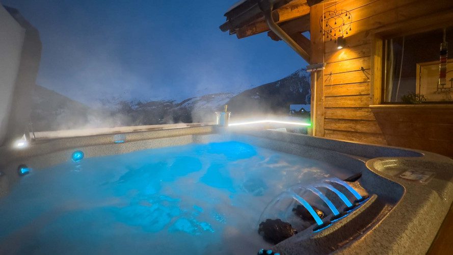 Steaming outdoor hot tub at Apartment La Cascade with illuminated blue water, wooden chalet exterior and snowy mountain views at dusk.