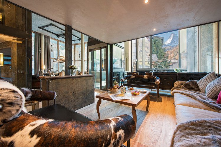 Comfortable lounge in the Backstage Luxury Loft with plush sofas, fur throws, a coffee table and large windows framing an alpine mountain view.