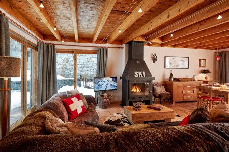 Open-plan lounge at Chalet Cest La Vie with plush seating, wood-burning fireplace, TV, dining area and exposed timber beams.