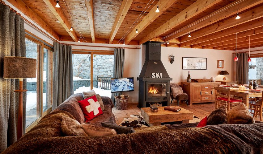 Open-plan lounge at Chalet Cest La Vie with plush seating, wood-burning fireplace, TV, dining area and exposed timber beams.