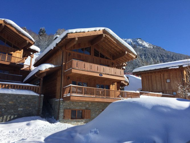 Exterior of Chalet Estournel in the snow with wooden alpine architecture, surrounding chalets and a clear blue sky.