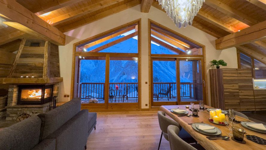 Open-plan lounge and dining area at Chalet Maison d'Etre with a fireplace, large picture windows and snowy mountain views.
