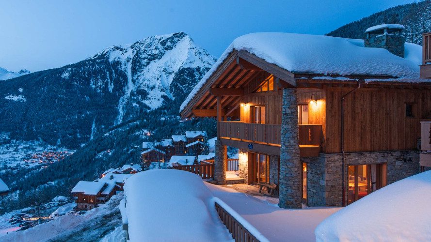 Exterior of Chalet Margaux at night with warm chalet lights, snow-covered surroundings, and alpine mountain views.