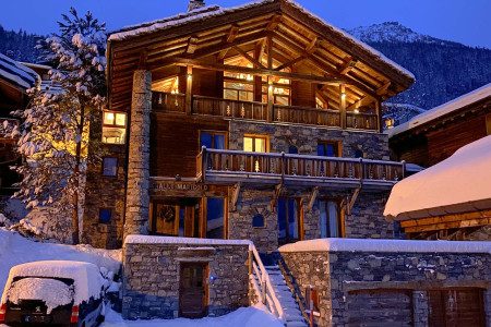 Dusk exterior of Chalet Marigold with warm interior lights glowing through large windows, surrounded by snow-covered alpine buildings.