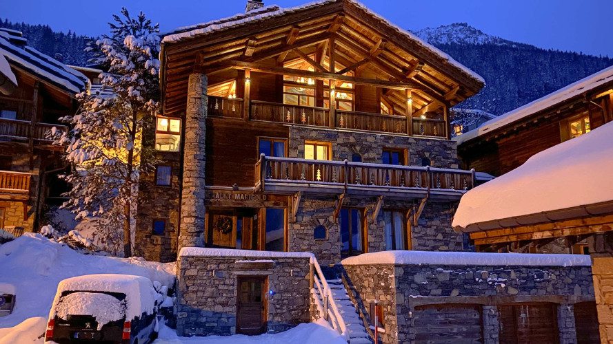 Dusk exterior of Chalet Marigold with warm interior lights glowing through large windows, surrounded by snow-covered alpine buildings.