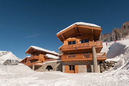 Exterior of Chalet Rayas surrounded by deep snow, with timber balconies, stone walls and a clear blue winter sky.
