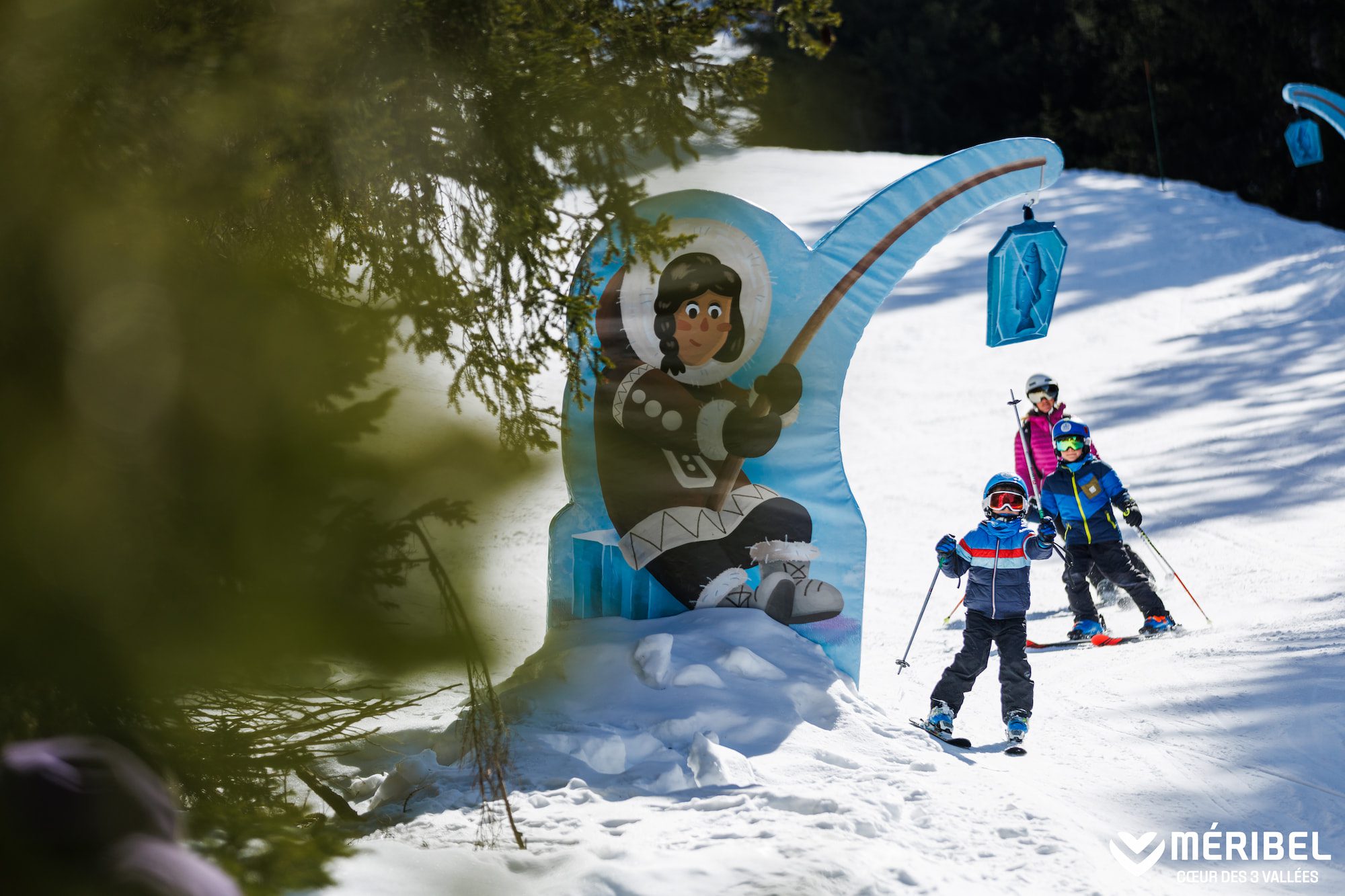 Children skiing in Méribel beside a playful Eskimo fishing-themed slope decoration on a snowy family ski run.