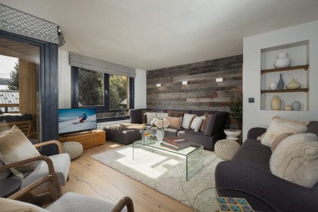 Main living room at Chalet La Muse with dark sofas, a glass coffee table, TV, wood feature wall and large windows.