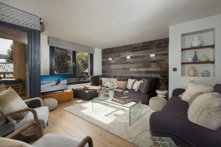 Main living room at Chalet La Muse with dark sofas, a glass coffee table, TV, wood feature wall and large windows.